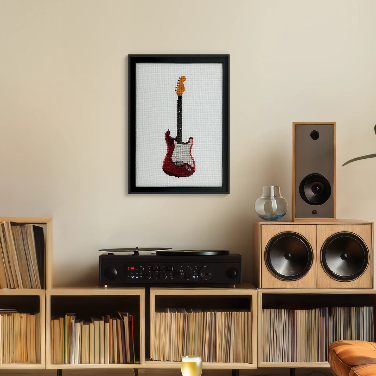 cross stitch of a red fender stratocaster guitar on white fabric in a black frame hung up on a wall above a record player