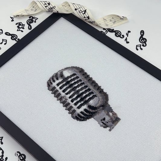 Cross-stitched retro microphone design on white fabric in a black frame.