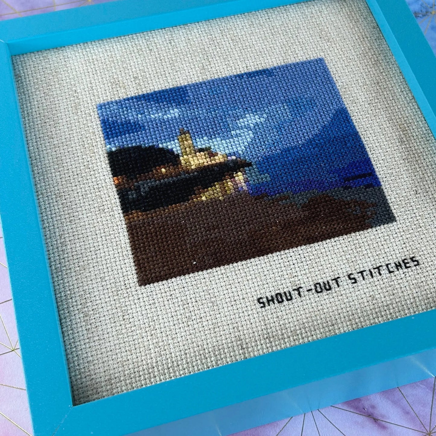 cross stitch on rustic aida fabric of church, trees, beach and sky in the evening in gijon, asturias, spain. In a blue square frame.