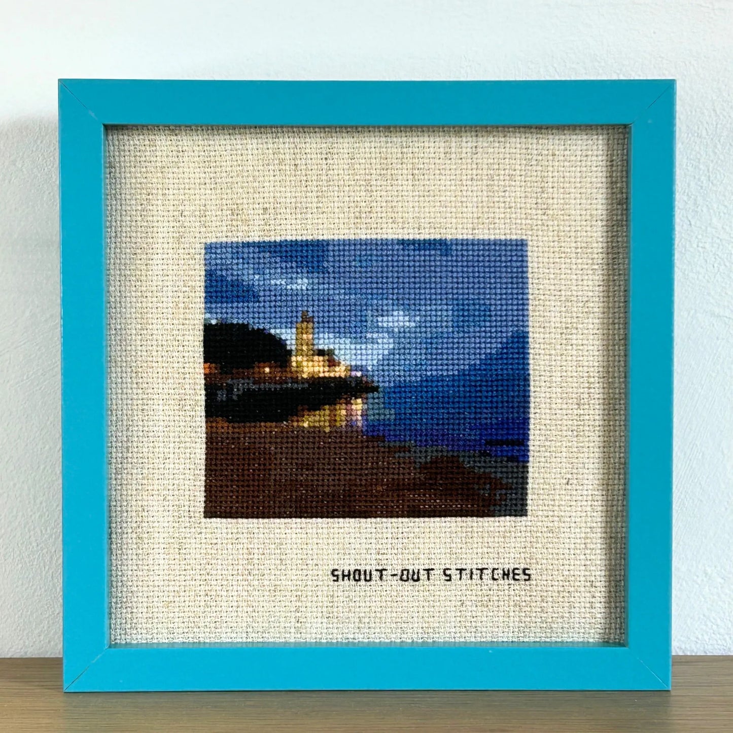 Cross stitch of an evening view showing a church, beach, trees and the sky in Gijon, Asturias, Spain. Sitched on rustic Aida fabric in a square blue frame.