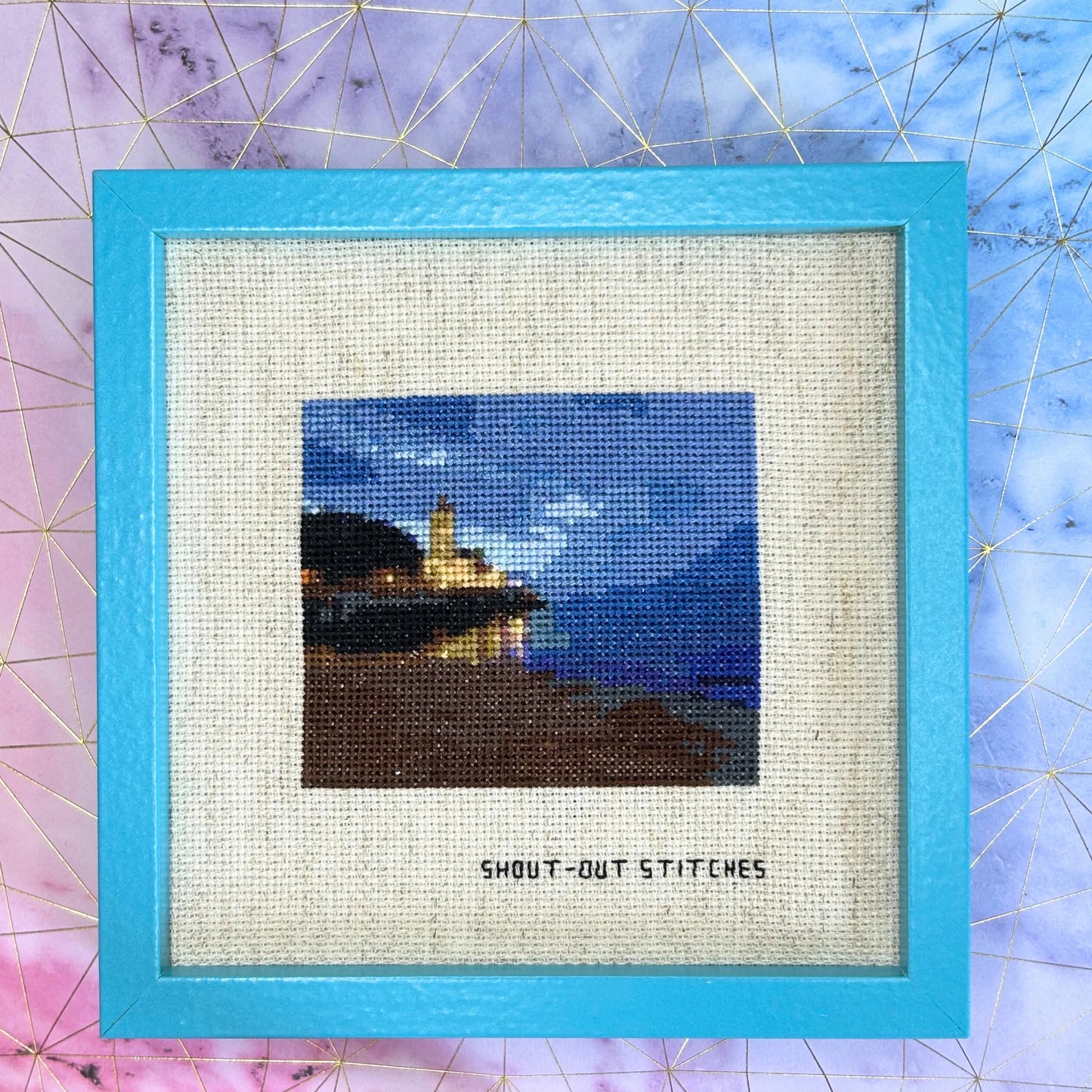 Cross stitch of an evening view of a church, sea, beach and sky of Gijon, Asturias, Spain. Stitched on Aida fabric in a blue square frame.