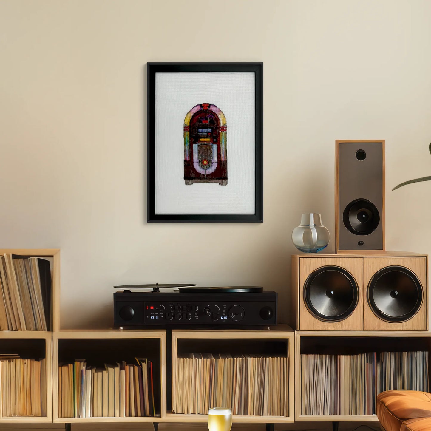 Cross stitch of a retro jukebox on white fabric in a black frame hung up on a wall above a record player