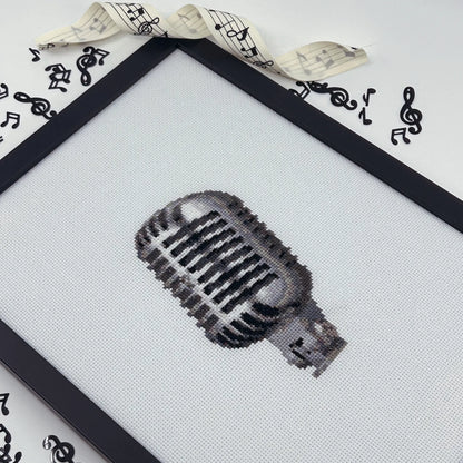 Cross-stitched retro microphone design on white fabric in a black frame.